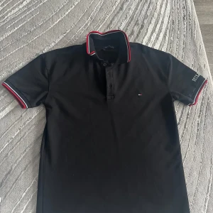Svart piké från Tommy Hilfiger  - Svart pikétröja från Tommy Hilfiger med  logga på ärmen och liten flagga på bröstet. den är med knappar vid halsen. Perfekt för en stilren look.