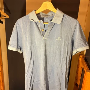 Ljusblå pikétröja från Gant - Säljer en klassisk ljusblå pikétröja från Gant med korta ärmar och krage. Tröjan har två knappar framtill och diskret Gant-logga broderad på bröstet. Perfekt för en chill och stilren look. Använd 1-2 gånger.