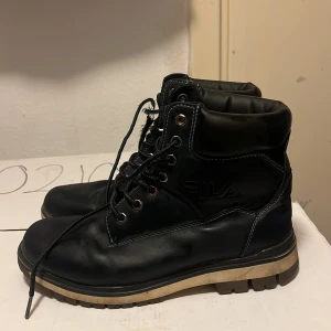 Svarta boots från Fila i skinn - Säljer ett par svarta boots från Fila med robust sula och vadderad kant runt ankeln. Skorna har snörning med metallhål och är tillverkade i skinn med kontrastsömmar. Perfekta för dig som vill ha en stilren och tålig sko till höst och vinter.