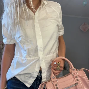 Vit kortärmad skjorta från Acne Jeans - Vit skjorta från Acne Jeans i klassisk design med korta ärmar och knappar framtill. Skjortan är tillverkad i bomull och har en avslappnad passform med lätt rynkade detaljer vid axlarna. Perfekt för en clean och stilren look.