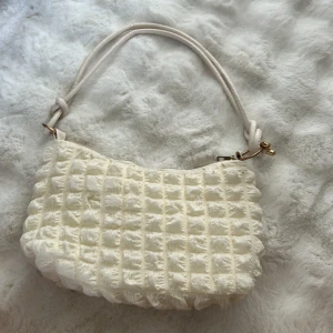 Vit quiltad axelväska med struktur - Superfin axelväska i fluffigt, quiltat tyg med rutmönster. Väskan är vit och har ett mjukt, strukturerat material som känns lyxigt. Handtaget är i ljus syntet med snygga knutdetaljer och guldfärgade metalldelar. Stängs med dragkedja och har ett rymligt innerfack.