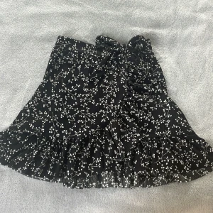 Svart blommig volangkjol! - Supersöt svart kortkjol från Trendyol med vitt blommönster. Kjolen har volangkant nertill och en snygg knytning i midjan som man kan ändra själv. Kjolen är storlek Xs men passar även mig som är S i vanliga fall!❤️