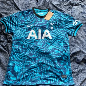 Tottenham Hotspur Son 7 Nike bortatröja - Säljer en Tottenham Hotspur bortatröja från Nike med Son 7 på ryggen. Tröjan är blå med mörkblå och turkosa mönster, har korta ärmar och är tillverkad i Dri-FIT-material. Stort AIA-tryck på bröstet och Spurs-logga broderad. Perfekt för dig som älskar fotboll och Spurs. Helt i nyskick är tröjan lappar finns kvar. Storlek är L och passar dom som är 175-180cm lång. Vid minsta fundering eller om du vill ha mer bilder på tröjan hör av dig.