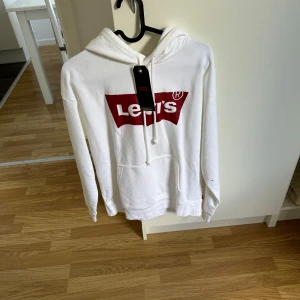 Vit hoodie från Levi's med logga - Vit hoodie från Levi's med stor röd logga framtill. Klassisk modell med huva, snörning och känguruficka. Mjuk bomullskänsla och långärmad passform, perfekt för en chill och stilren look. Aldrig använd 