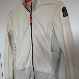 Vit och grå overshirt från Parajumpers - Snygg vit och ljusgrå overshirt från Parajumpers med dragkedja framtill och ribbade muddar. Jackan har en cool patch på ärmen och orange detalj vid dragkedjan. Perfekt för dig som gillar stilren och sportig look.