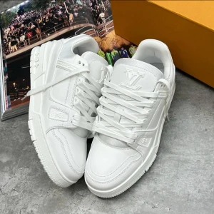 LV Trainer Sneaker - Snygga helvita Louis Vuitton sneakers med LV-logga på plösen och detaljerade sömmar. Skorna har en chunky sula, rund tå och är tillverkade i skinn med textilinslag. Klassisk snörning och diskreta logodetaljer på sidan och sulan. Perfekta för dig som gillar clean och lyxig streetstyle.