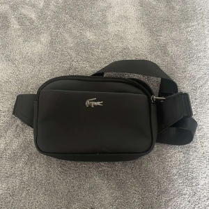 Svart axelväska från Lacoste - Snygg svart axelväska från Lacoste med klassisk krokodil-logga i silver framtill. Väskan har justerbart band, dragkedja och ett rymligt huvudfack med innerficka. Perfekt för att bära det viktigaste och passar till en clean streetstyle.
