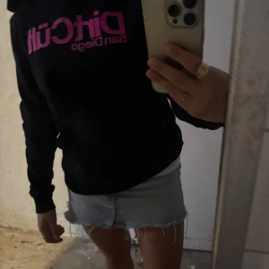 Svart DirtCült hoodie med rosa tryck - Svart hoodie från DirtCült med stor rosa logga och texten 'San Diego' på bröstet. Klassisk känguruficka framtill och huva. Perfekt för dig som gillar streetstyle och vill sticka ut med en cool kontrast mellan svart och neonrosa.