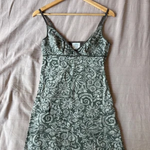 Grön blommig kortklänning med smala axelband - Supersöt grön kortklänning med blommigt mönster och smala axelband. Klänningen har v-ringad urringning och figurnära passform upptill. Perfekt för varma dagar när du vill känna dig både chill och snygg.