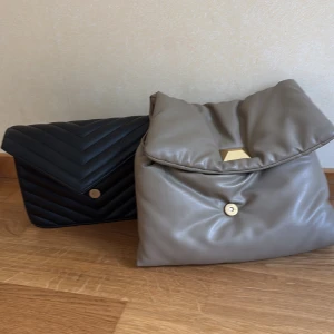 Två quiltade axelväskor med kedja - Säljer två snygga axelväskor i quiltad design med kedjeremmar. Den ena är svart med hjärtdetalj på baksidan och guldknapp, den andra är beige med stor guldspänne och mjuk, puffig look. Båda i skinnimitation och perfekta för att lyfta din outfit.