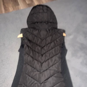 Svart dunväst med huva från Zara - Svart dunväst från Zara  Västen har en stor huva och är perfekt för lager-på-lager-stil. Passformen är normal och materialet är mjukt och värmande syntet.