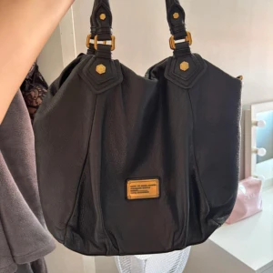 Svart axelväska Marc by Marc Jacobs - Snygg svart axelväska i skinn från Marc by Marc Jacobs med guldiga metalldetaljer och logoplatta framtill. Väskan har dubbla handtag och ett rymligt foder med grafiskt tryck. Perfekt för dig som vill ha en klassisk men cool väska med plats för allt du behöver.