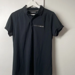 Svart pikétröja från Sail Racing - Svart pikétröja från Sail Racing i klassisk modell med krage och korta ärmar. Tröjan har diskret logga på bröstet och en liten logga i nacken. Tillverkad i mjuk bomull, perfekt för en clean och sportig stil.