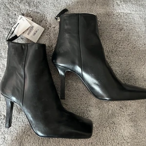 ZARA skinnstövletter med klack HELT NY - Stilrena svarta stövletter från Zara.Modellen har fyrkantig tå, smal hög klack på 5 cm och dragkedja bak. Perfekta för dig som gillar minimalistisk och trendig look. Snygga till allt. HELT NY, oanvänd och ursp. pris låg på 1295 kr. 