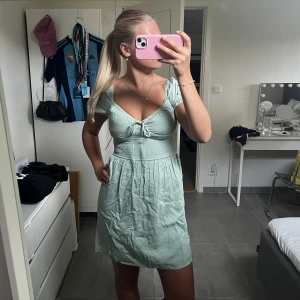 Ljusgrön kort klänning från Bik Bok - Supersöt ljusgrön kort klänning från Bik Bok med smockad midja, puffärmar och söt knytning framtill. Klänningen har v-ringning och är gjord i ett lätt bomullstyg, perfekt för varma dagar. Passformen är normal och den har en flowy kjol.