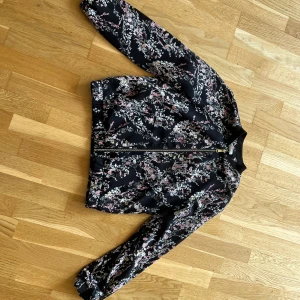 Blommig bomberjacka  - Snygg bomberjacka med svart bas och blommigt mönster i rosa, vitt och gult. Jackan har guldig dragkedja och en ribbad krage. En perfekt lite tunnare jacka. Det är bara att fråga om du undrar något! 😊