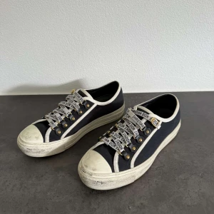 Svarta Dior sneakers med vita detaljer - Snygga svarta sneakers från Dior med vita kanter och coola snören med texten 'Christian Dior J'Adior'. Skorna har platt sula, rund tå och guldfärgade metalldetaljer vid snörningen. Tillverkade i canvas för en trendig och avslappnad look.