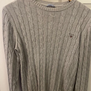 Grå kabelstickad tröja från Gant - Kabelstickad grå tröja från Gant med rund halsringning och diskret logga broderad på bröstet. Långärmad och i klassisk, tidlös design som är perfekt för lager-på-lager. Mjuk och skön känsla, passar dig som gillar stilrena outfits! Hör av er för mer frågor 