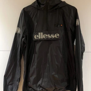 Svart vindjacka från Ellesse - Svart vindjacka från Ellesse med stor logga framtill och huva med snörning. Jackan har lång ärm, dragkedja upptill och en stor ficka på magen. Perfekt för dig som gillar sportig och clean stil.