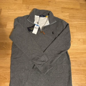 Grå half zip tröja från Polo Ralph Lauren - Snygg grå långärmad tröja från Polo Ralph Lauren med klassisk broderad logga på bröstet. Tröjan har en half zip-dragkedja med läderdetalj och hög krage. Perfekt för lager-på-lager och chill stil. Tillverkad i mjuk bomull för extra komfort.
