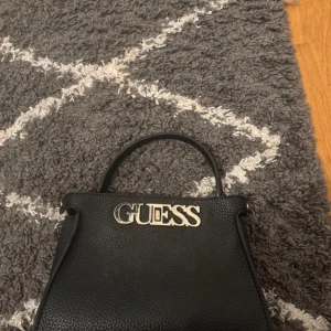 Svart handväska från Guess - Snygg svart handväska från Guess med stort metallogo framtill. Väskan är i strukturerat skinn och har ett stilrent, klassiskt utseende med ett handtag upptill. Perfekt för dig som vill ha en trendig och tidlös accessoar.