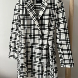 Blazerklänning med dubbelknäppning  - Den här klänningen är en stilren och modern blazerklänning från märket Bershka. Blazerklänning med dubbelknäppning (double-breasted) framtill. Färg och mönster: Vit bas med ett klassiskt svart rutigt mönster (typiskt tartan- eller rutigt inspirerat). Krage: Klassisk kavajkrage med slag. Ärmar: Långa ärmar, raka. Stil: Den har ett strukturerat, skräddarsytt utseende som gör att den passar både som klänning och som lång blazer/kavaj.