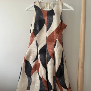 Mönstrad kortklänning från H&M - Snygg ärmlös kortklänning från H&M med grafiskt mönster i svart, beige och rostbrunt. Klänningen har rund halsringning, markerad midja och dragkedja i ryggen. Materialet är strukturerat.