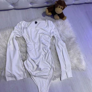 Vit långärmad omlott bodysuit - Använd en gång, gick ner i vilkt så Säljer en vit långärmad bodysuit  med snygg omlott-effekt och rynkade detaljer längs sidorna. Den har en djup, något v-ringad halsringning och är tillverkad i mjukt stretchigt material som sitter skönt på kroppen.