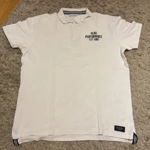 Vit Peak Performance pikétröja XL - Snygg vit piké från Peak Performance i storlek XL. Klassisk design med korta ärmar, krage och knappar framtill. Diskret logga på bröstet och coola randiga detaljer vid slitsarna nertill. Perfekt för en clean och sportig look.