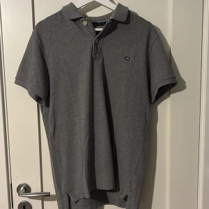 Grå pikétröja från Lexington - Klassisk grå pikétröja från Lexington med liten logga på bröstet. Tröjan har krage, knappar vid halsen och korta ärmar. Tillverkad i mjuk bomull som känns skön mot huden. Perfekt för en clean och avslappnad stil.