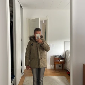 Beige parkasjacka med fuskpäls från Lindex - Snygg beige parkasjacka från Lindex med huva och fuskpäls. Jackan har broderade patches på bröstet och ärmarna, stora fickor framtill och dragkedja med knappslå. Perfekt för kalla dagar och riktigt skön att bära.