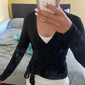 Svart omlottkofta med knytband - Snygg svart omlottkofta med långa ärmar och knytband i midjan. Koftan har v-ringad halsringning och sitter snyggt figurnära. Perfekt att styla över ett linne eller t-shirt för en clean look.