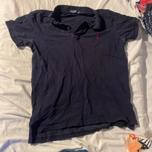 Svart pikétröja från Polo Ralph Lauren - Klassisk svart pikétröja från Polo Ralph Lauren med korta ärmar och krage. Tröjan har en röd broderad logga på bröstet och är gjord i mjuk bomull. Perfekt för en clean och stilren look.