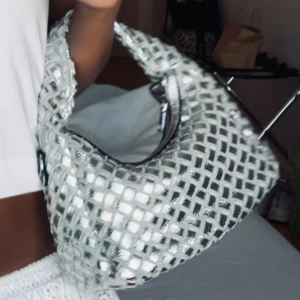 Silvrig axelväska med flätad look - Säljer en unik silvrig axelväska med flätad design och glittrig yta. Väskan har ett kort handtag och en avtagbar axelrem i matchande silver. Stängs med dragkedja och har en cool, modern form som sticker ut.
