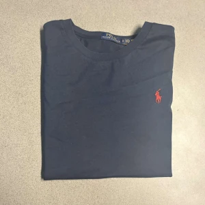 Mörkblå t-shirt från Polo Ralph Lauren - Klassisk mörkblå t-shirt från Polo Ralph Lauren med rund hals och korta ärmar. T-shirten har den ikoniska röda broderade loggan på bröstet och är gjord i mjuk bomull. Perfekt basic-plagg med snygg passform.