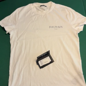 Vit Balmain Paris t-shirt i bomull - Snygg vit t-shirt från Balmain Paris med diskret logga tryckt på bröstet. Klassisk rund halsringning och korta ärmar. Tillverkad i mjuk bomull för en skön känsla och clean look. Perfekt för dig som gillar stilrena designer och exklusiva märken.