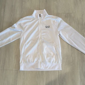 Vit track jacket från EA7 Emporio Armani - Snygg vit track jacket från EA7 Emporio Armani med dragkedja framtill och höga krage. Jackan har två fickor med dragkedja och svart logga på bröstet. Perfekt för en sportig och clean look.