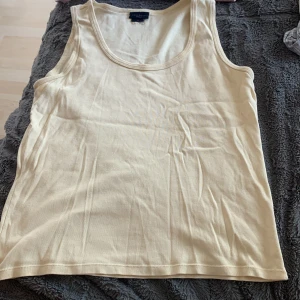 Beige linnetopp från Gant - Snygg och enkel beige linnetopp från Gant i klassisk modell. Toppen är ärmlös med rund halsringning och tillverkad i mjuk bomull, perfekt för varma dagar eller som lager under skjorta. Stilren och lättmatchad till det mesta.