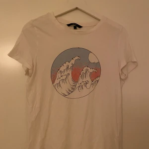 Vit T-shirt  - Vit t-shirt från Vero Moda med korta ärmar och rund hals. Framsidan har ett stort tryck med vågor i blått och rosa tone. Skön passform och tillverkad i mjuk bomull.