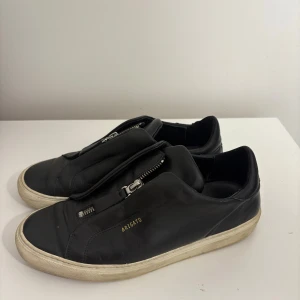 Svarta sneakers från Axel Arigato - Svarta sneakers i skinn från Axel Arigato med cool dragkedjedetalj på ovansidan och vit sula. Minimalistisk design med guldfärgad logga på sidan. Perfekta för dig som gillar stilrena och moderna skor med edge.