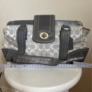 Coach grå handväska med logomönster - Snygg handväska från Coach i grått och silver med klassiskt logomönster. Väskan har detaljer i skinn, robusta handtag och praktisk dragkedja. Perfekt storlek för vardagens essentials och har en stilren look med metallspänne framtill.