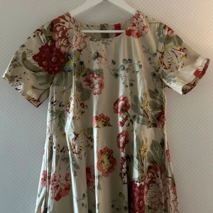 Blommig långklänning från Lina Woddlán - Superfin långklänning från Lina Woddlán i ljusbeige med stora blommor i rött, rosa, grönt och blått. Klänningen har korta ärmar, rund halsringning och ett flowy fall.