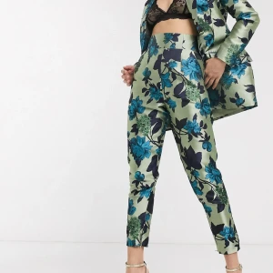 Blommiga kostymbyxor från ASOS - Snygga högmidjade kostymbyxor från ASOS i ljusgrön satin med blått och mörkblått blommönster. Byxorna har croppad längd och smal passform, perfekta för dig som vill sticka ut med en färgstark look.