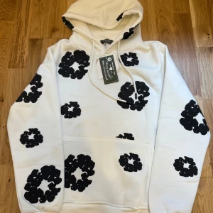 Vit hoodie med svarta blommor Denim Tears - Vit hoodie från Denim Tears med stora svarta blommiga tryck över hela tröjan. Tröjan har huva med snörning och en klassisk känguruficka framtill. Perfekt för dig som vill sticka ut med en cool och unik stil.