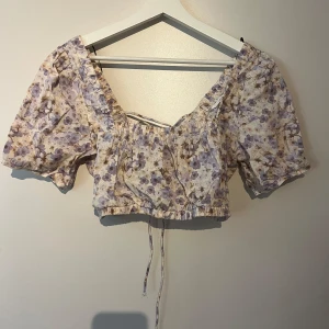 Blommig croppad blus från H&M - Superfin croppad blus från H&M i linnemix med lila och beige blommönster. Puffiga korta ärmar, v-ringning och snygg snörning i ryggen. Perfekt för vår och sommar när du vill ha en söt och trendig look.Den är aldrig använd endast provad men saknar prislapp