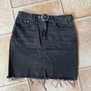 Mörkgrå jeanskjol med bälte och fransad kant - Mörkgrå jeanskjol med snyggt fransad nederkant och tillhörande bälte med rund metallspänne. Klassisk femficksmodell med dragkedja framtill och bakfickor. Perfekt för dig som gillar en edgy och avslappnad stil.