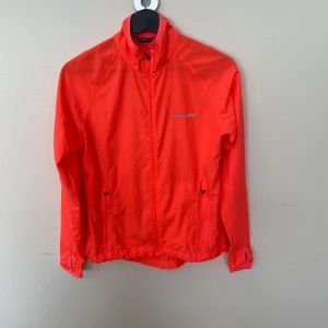 Neon orange vindjacka från SOC - Snygg neonorange vindjacka från SOC med hel dragkedja framtill och hög krage. Jackan har långa ärmar, två fickor med dragkedja och reflekterande detalj på ryggen. Använd 1 gång.