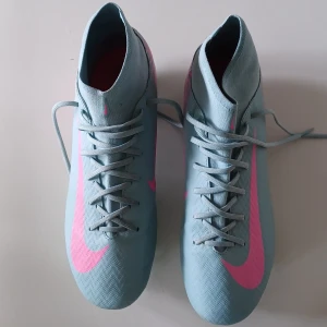 Nike Mercurial blå/rosa fotbollsskor - Snygga Nike Mercurial fotbollsskor i ljusblått med rosa detaljer och stora Swoosh-loggor. Skorna har snörning, högre strumpliknande skaft och mönstrad ovandel i syntetmaterial. Perfekta för dig som vill sticka ut på planen. Priset kan diskuteras vid snabbaffär. Minst 650.