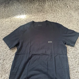 Svart t-shirt från BOSS - En stilren svart t-shirt från BOSS med diskret logga på bröstet. Klassisk rund halsringning och korta ärmar. Perfekt för en clean och enkel look. Tillverkad i mjuk bomull som känns skön mot huden.
