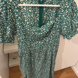 Grön blommig klänning med puffärm - Supersöt grön klänning med vitt blommönster och puffiga korta ärmar. Klänningen har en lätt draperad halsringning och stängs med dragkedja bak. Perfekt för sommarens alla tillfällen!